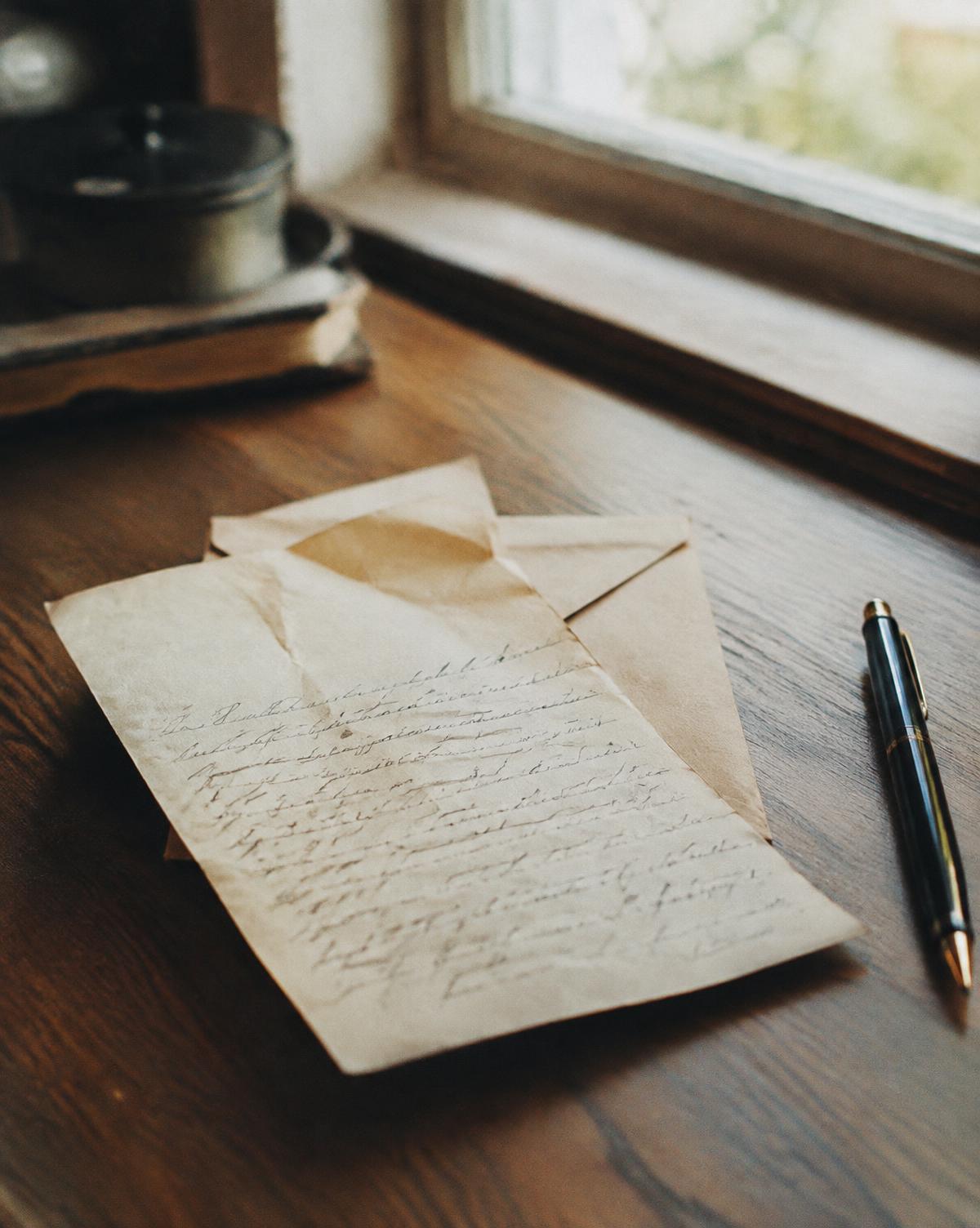 Carta manuscrita y un sobre abierto sobre un escritorio de madera junto a una ventana, con una pluma al lado