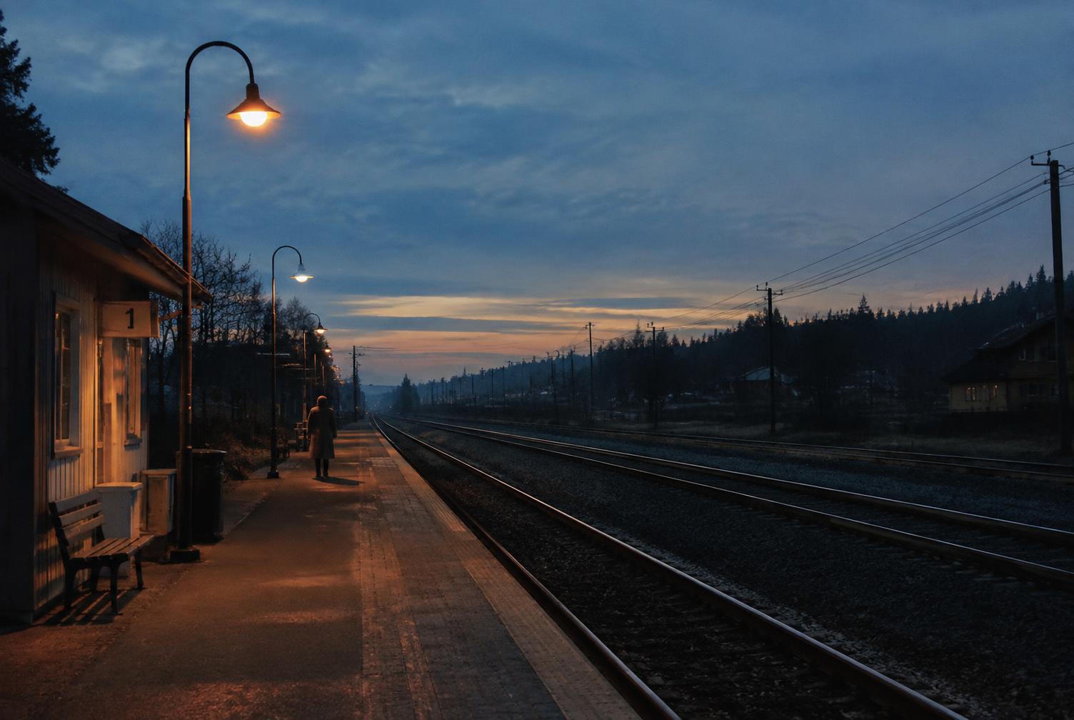 Andén de tren vacío al anochecer, con farolas encendidas y una figura lejana esperando